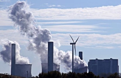 The wide, cylindrical columns of a large coal power plant throw clouds of white fumes into the air, while in the foreground, a lone wind turbine rises high above the tops of trees.