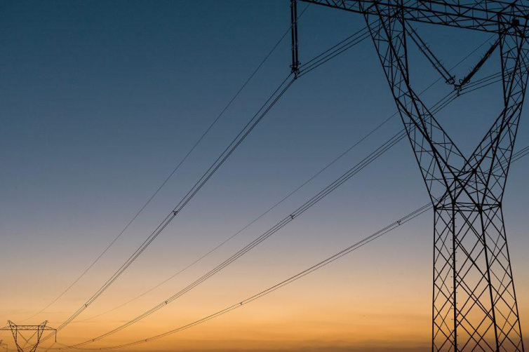 Sky at dusk with an electrical tower in the foreground joined to another that is in the background by electrical wires.