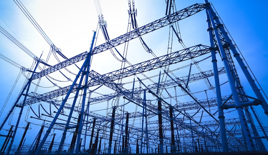 An electrical transformer grid, with many poles and wires, beneath a blue sky and white light overhead.