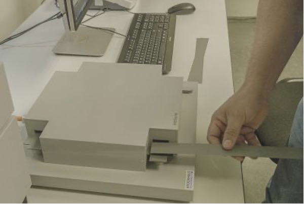 An individual is interacting with a large, intricate mechanical device resting on a table in an indoor setting. In the background, various electronic devices, including a monitor and a keyboard, are visible. A hand is visible, grasping a portion of the mechanical device. The mechanical device is grey and consists of complex layers and components. It is placed on a white table, contrasting with its grey color.