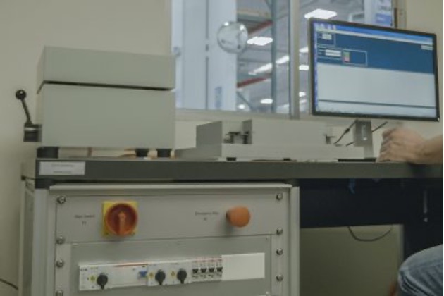 A person is operating a large and complex mechanical device inside a room. The grey machine has various components, such as control knobs, indicators, and a button. There is also a computer monitor displaying a blue screen on the table. The person’s arm is visible, and in the background, blurred objects and electronic devices hint at a laboratory or workspace environment.