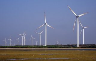 Under a cloudless sky there are numerous wind turbines with three immense blades per turbine in use along a vast marsh.