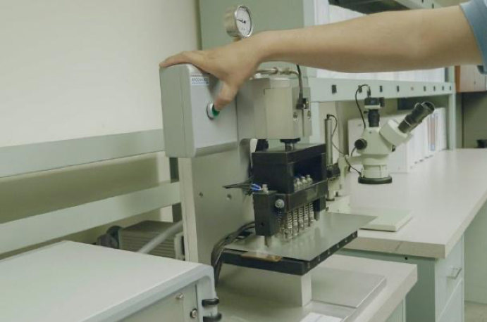 A worker’s hand can be seen pressing a green button on a machine that has several cylindrical tubes pressed against a flat steel metal part about the size of a book, behind which a microscope and a long bare counter with drawers.