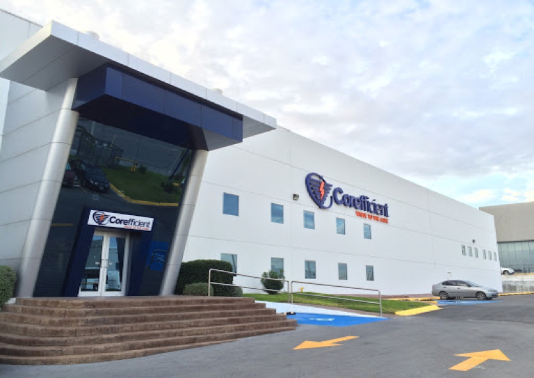 An outside view of the Corefficient manufacturing plant: a blue glass entrance, a white building with the blue Corefficient logo, and the parking lot.