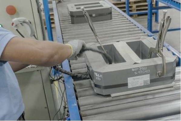 An industrial setting, specifically a production or assembly line where a person’s arm is visible, reaching out to assemble objects on a grey conveyor belt. Each electrical steel has white labels with printed text and barcodes for identification and tracking purposes. Blue metal structures in the background mounted on a post near the conveyor belt complete the scene. The overall atmosphere suggests manufacturing or assembly processes.