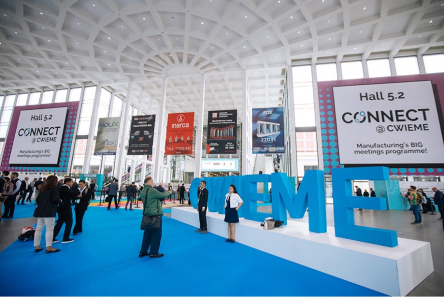Pictured is a large indoor area with a high ceiling, big windows, and an attractive modern design with white architectural shapes. Big blue letters at the center of the atrium spell “CWIEME.” Lots of people are talking and walking around the space. Banners advertising different companies are hanging from the ceiling.