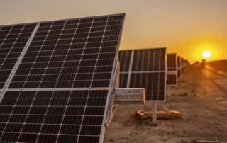 A long roll of silver and gray colored solar panels, stretching down a dusty road, into the background, towards a setting sun.