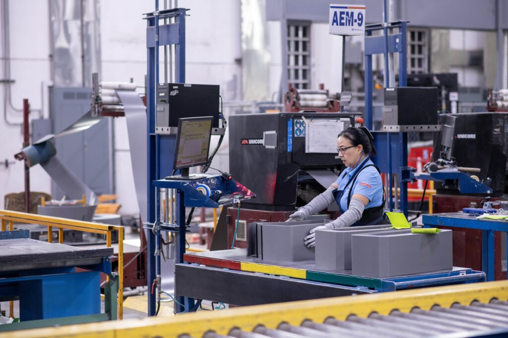 Corefficient facility worker assembling transformer cores in a manufacturing environment. The worker, wearing safety gear, handles components near industrial equipment labeled “Unicore.” Corefficient’s logo is visible in the lower right of the image.