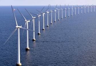A row of offshore wind turbines lined up in the ocean with clear blue skies and a ship in the background