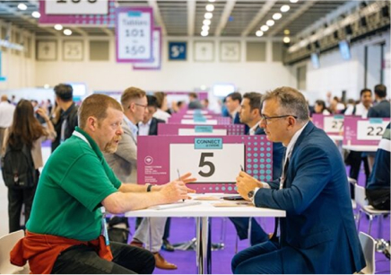Two people are engaged in a conversation at the CWIEME event, seated opposite each other at a table labeled “CONNECT 5.” The backdrop is a spacious exhibition hall bustling with similar pairs at tables, with booths and attendees in the background representing a professional trade show environment.