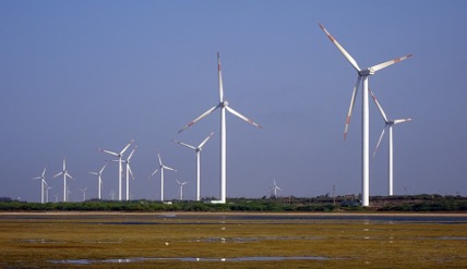 Under a cloudless sky there are numerous wind turbines with three immense blades per turbine in use along a vast marsh.