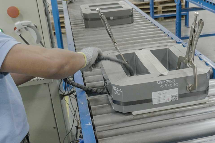 The arm and hand of a worker with nylon gloves and a short-sleeve blue shirt is working on a cable attached to an electric core which is a grey, metallic structure about half the size of a small suitcase that resembles a rectangle with wide walls and a hollow interior except for a bar running from one side of the rectangle to the other. This electric core is one of two on an assembly line in a factory.