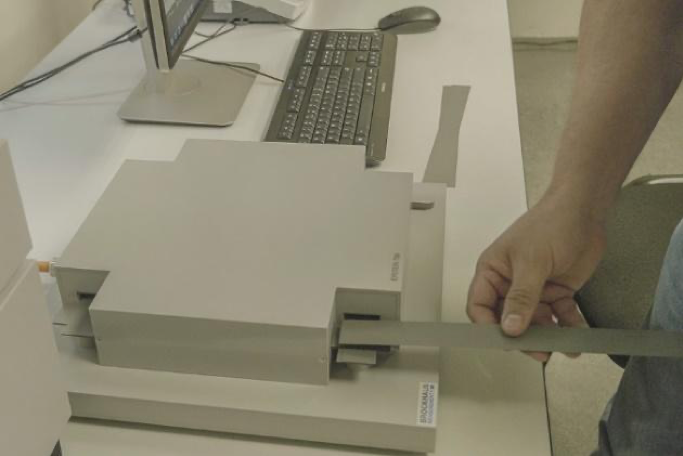 An image of a plain, off-white device laying on a table that looks like a “plus” sign into which the hand of a worker is inserting a slim, metal piece that resembles a ruler and a keyboard, mouse and computer screen behind the machine.