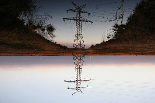 A large power line tower, with its reflection being beautifully mirrored in a nearby body of water